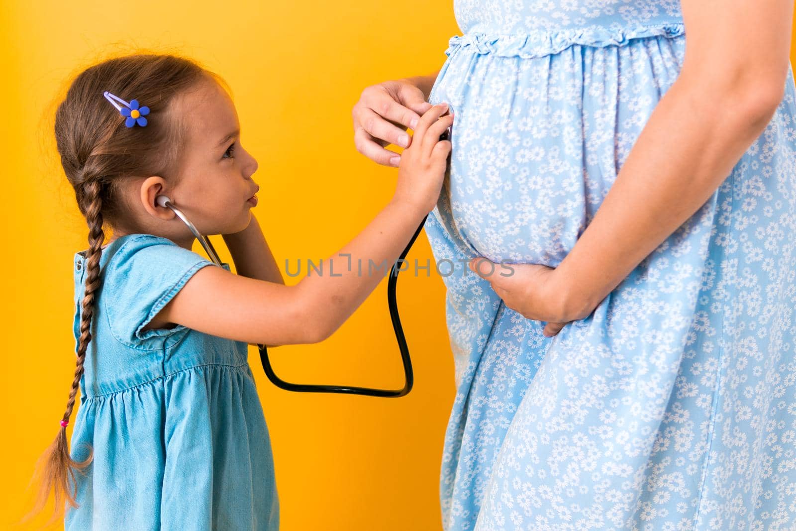 Motherhood, hot summer - croped portrait pregnant mother unrecognizable woman blue dress little Daughter girl sibling treat mom role play game stethoscope unborn big belly tummy on yellow background.