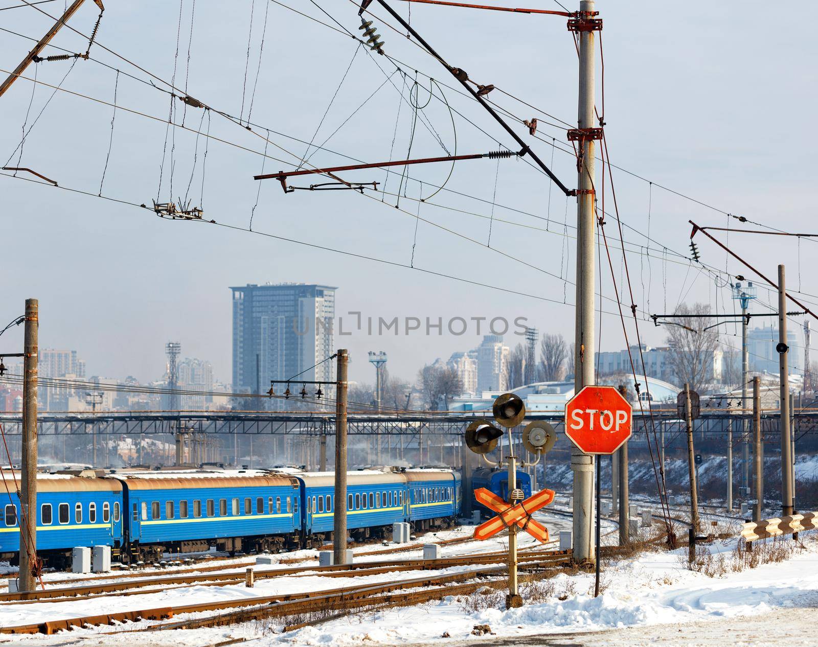Red stop sign at the railway crossing against the background of the railway station and buildings of the winter city. by Sergii