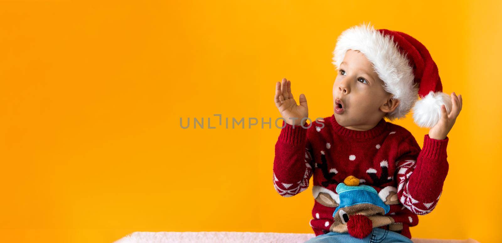 Portrait of happy smiling positive joyful positive preschool little boy in red warm santa hat showing thumb up on orange, yellow background. Winter, holiday, celebration, Christmas, New Year copy space.