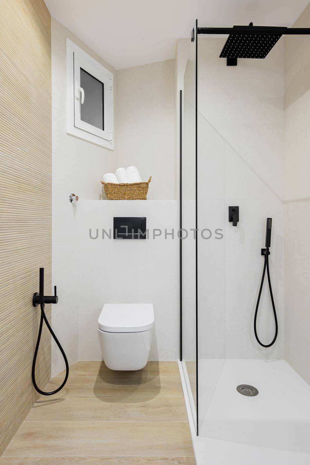 Interior of modern style bathroom in white and beige colors in refurbished apartment. Shower zone and toilet, with black faucets, towels and tiled floor and walls