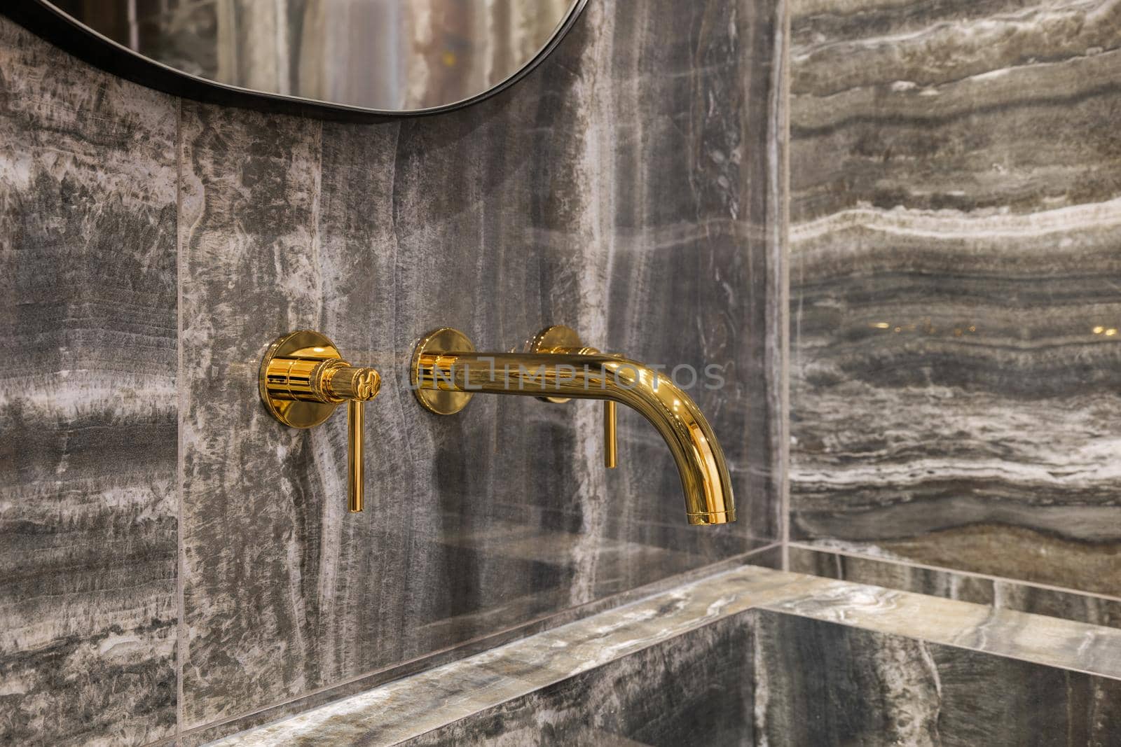 Close-up on modern golden tap in bathroom washbasin with marble wall tiles.