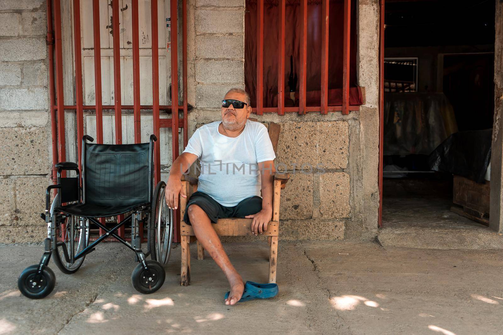 Disabled mature Latin man without a leg resting outside his house with dark glasses and his wheelchair by cfalvarez