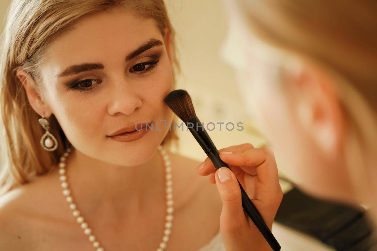 Make-up artist makes professional makeup of a young woman, a bride. The bride in a white dress, a pearl necklace and earrings. Make-up artist applies powder. by Matiunina