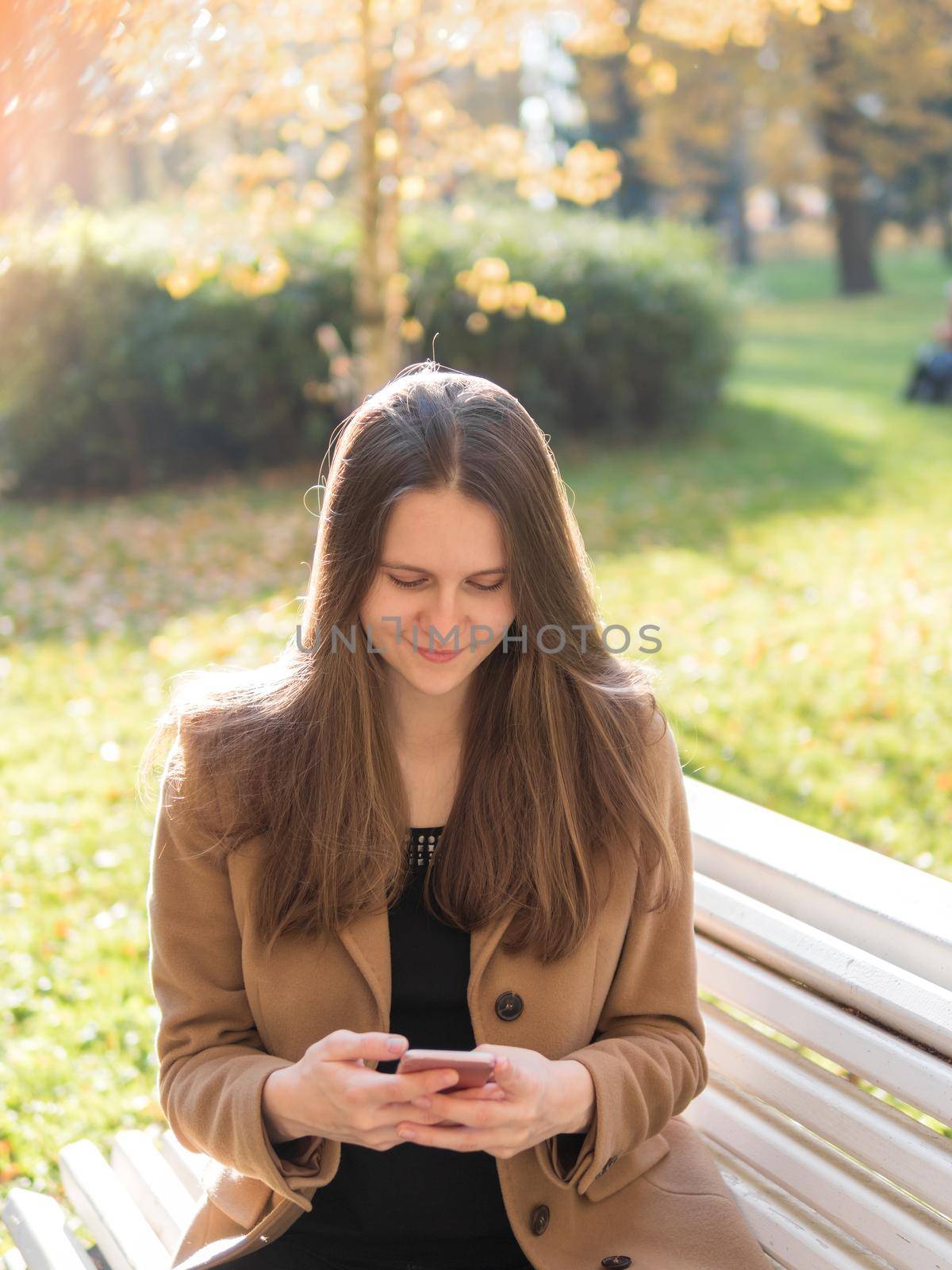 Beautiful teen girl sitting in the Park on a bench, holding a smartphone and chatting online on the Internet. Woman with long hair by NataBene