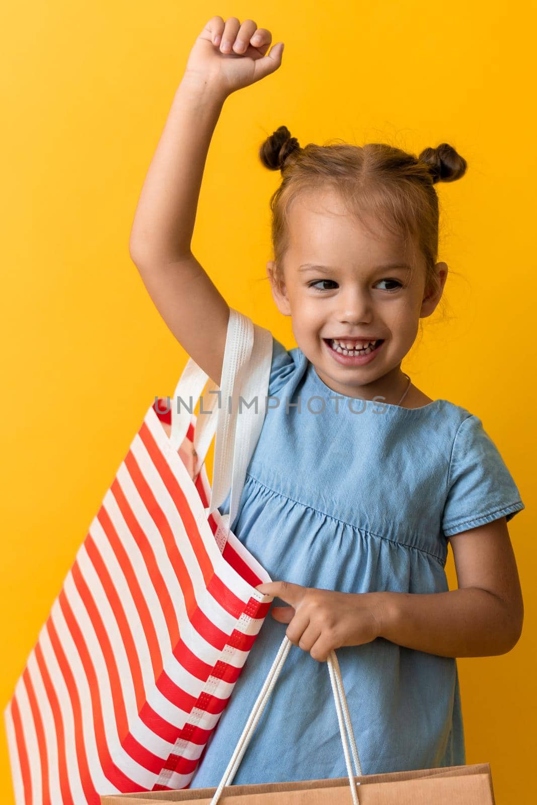 Portrait Caucasian Beautiful Happy Little Preschool Girl Smiling Cheerful And Holding Cardboard Bags Isolated On Orange Yellow Studio background. Happiness, Consumerism, Sale People shopping Concept by mytrykau