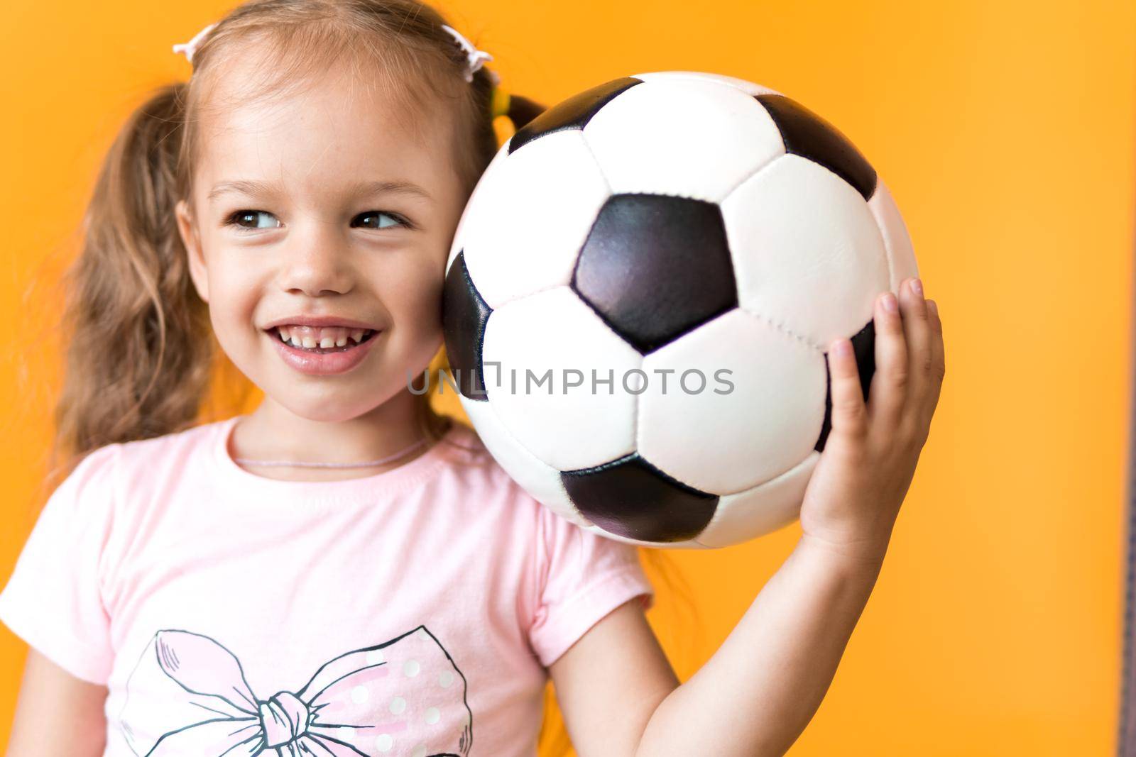 Authentic cute smiling preschool little girl with classic black and white soccer ball look at camera on yellow background. child play football in t-shirt and shorts. Sport, championship, team concept.