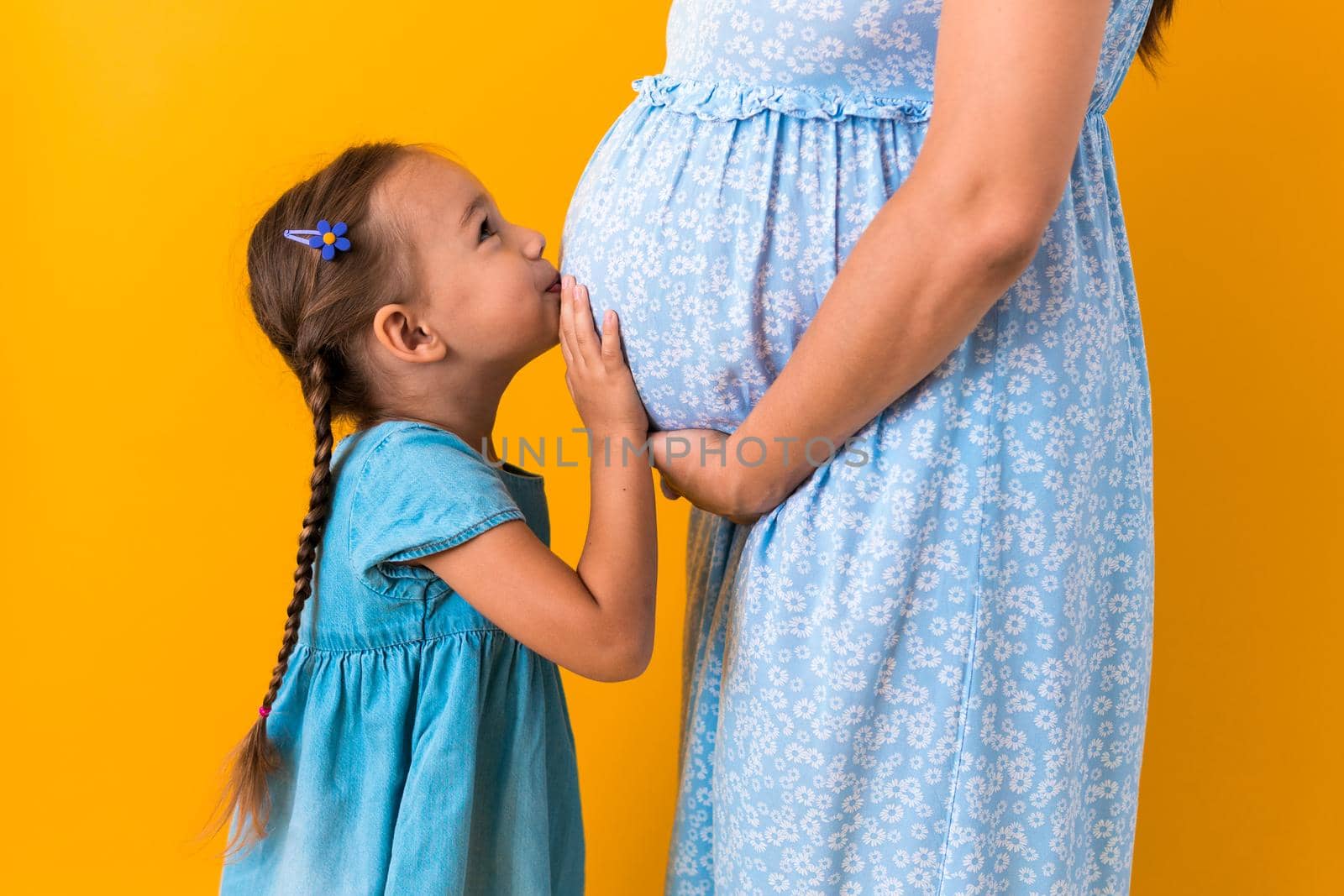 Motherhood, love, Childhood, pregnancy, hot summer - croped portrait pregnant unrecognizable mother woman blue dress little preschool Daughter girl sibling teddy bear hug mum belly on yellow background.