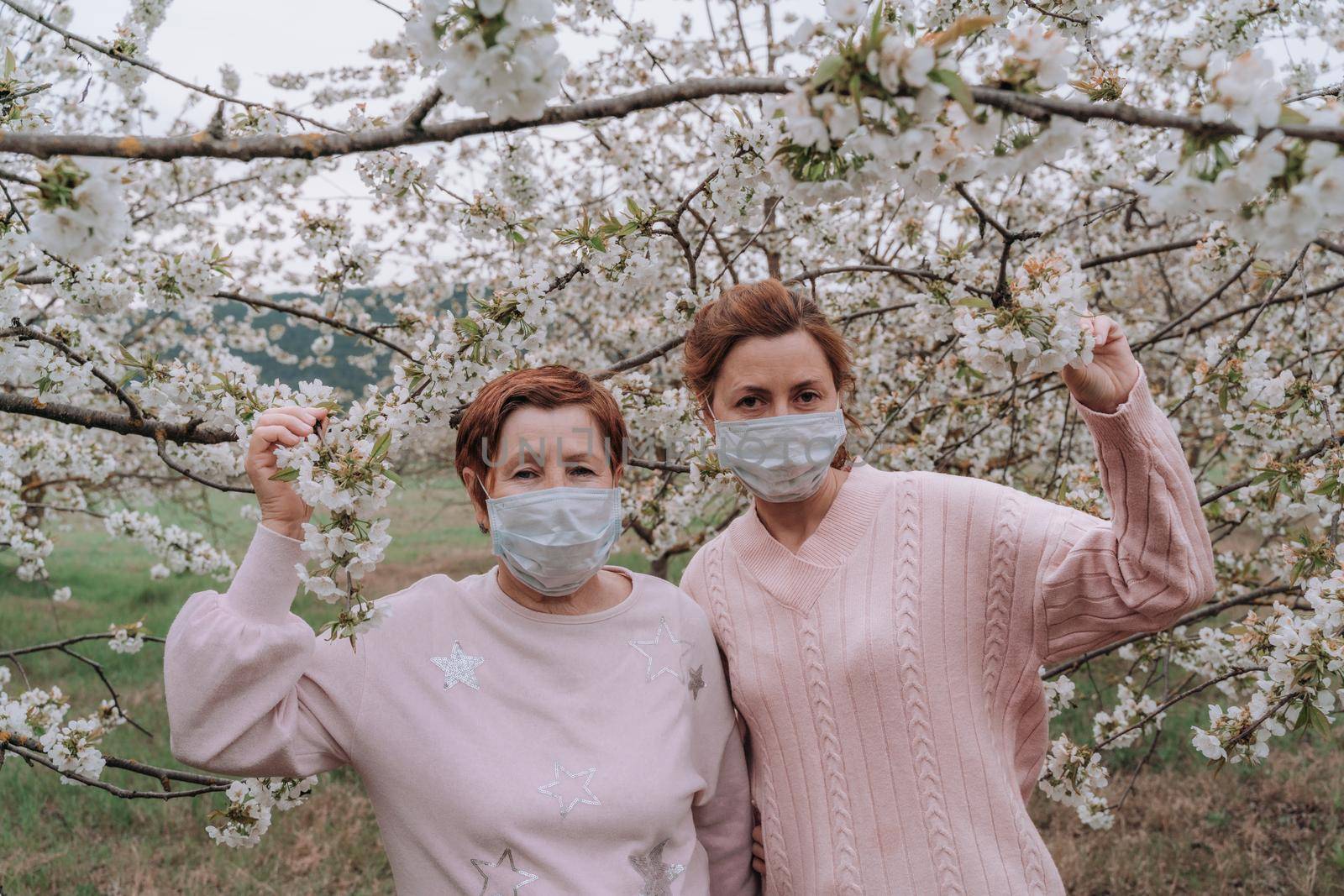 Mom and daughter in medical masks in pink sweaters are standing in a flowering cherry garden. Quarantine concept, next to the house. by Matiunina