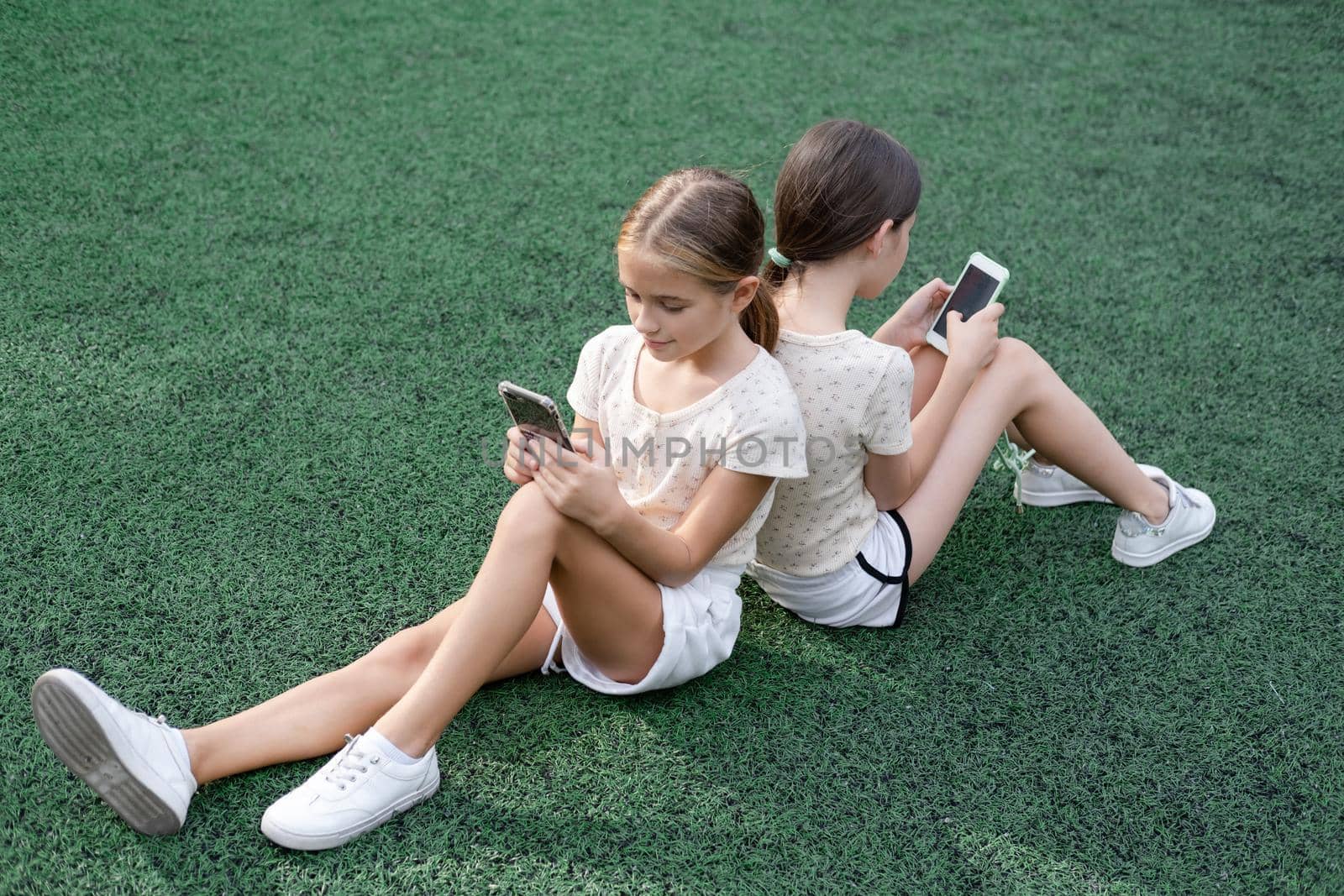 two pretty girls in identical clothes using phone on sports ground or athletic field. kids are surfing in internet. smartphone addiction. advanced, modern children in social networks by oliavesna