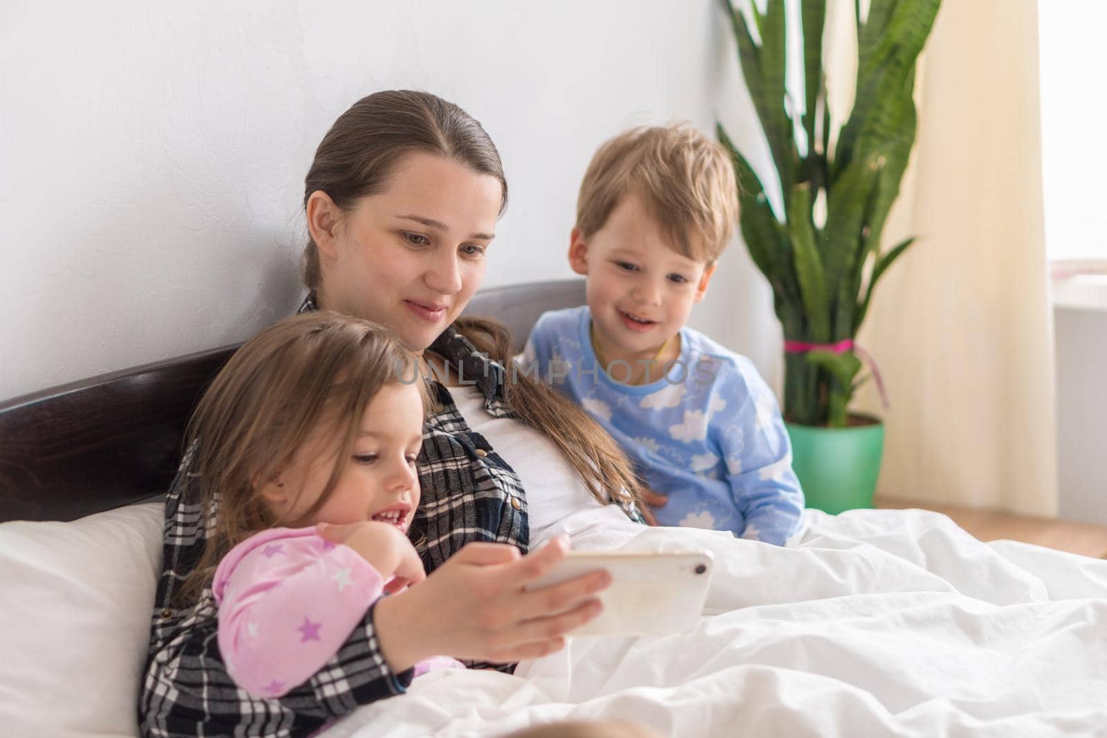 Happy Young Woman Mothefamilyr With 3-4 Aged Children Siblings Baby Boy And Girl Watching Smartphone Together Digital Tablet On White Bed. Mom With Daughter And Son Family Making Selfie In Pajamas