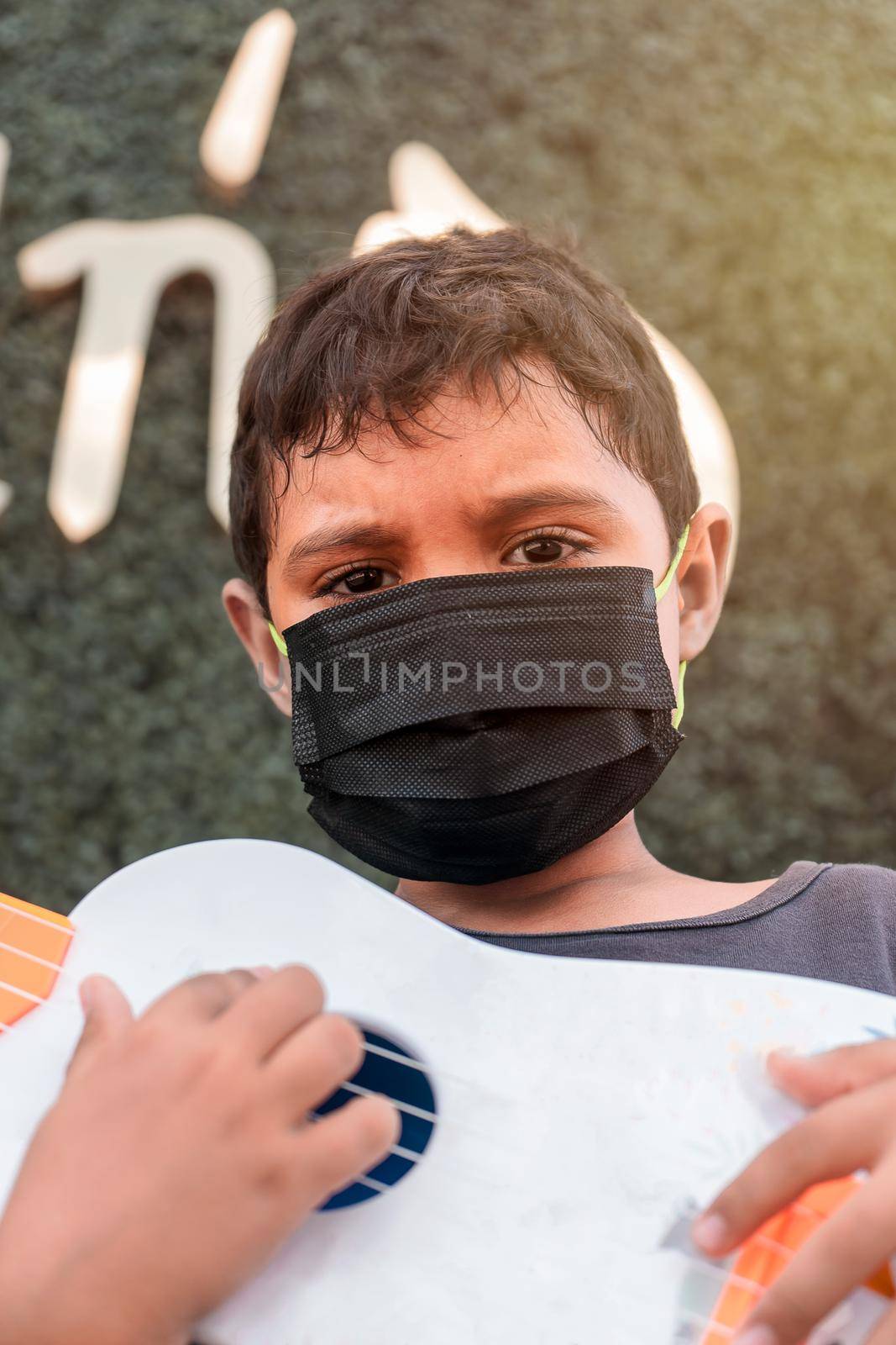 Vertical photo of a Latin boy with a medical mask outdoors looking at the camera and with a ukulele in his hands.