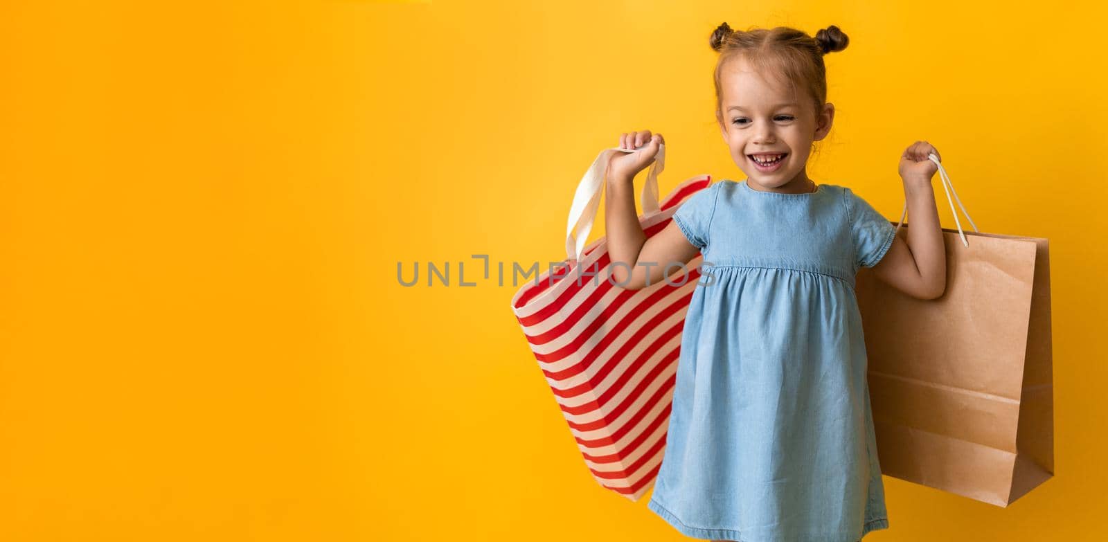 Banner Portrait Caucasian Beautiful Happy Little Preschool Girl Smiling Cheerful And Holding Cardboard Bags Isolated On Orange Yellow Background. Happiness, Consumerism, Sale People shopping Concept.