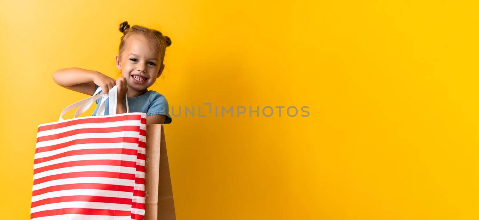 Banner Portrait Caucasian Beautiful Happy Little Preschool Girl Smiling Cheerful And Holding Cardboard Bags Isolated On Orange Yellow Background. Happiness, Consumerism, Sale People shopping Concept by mytrykau