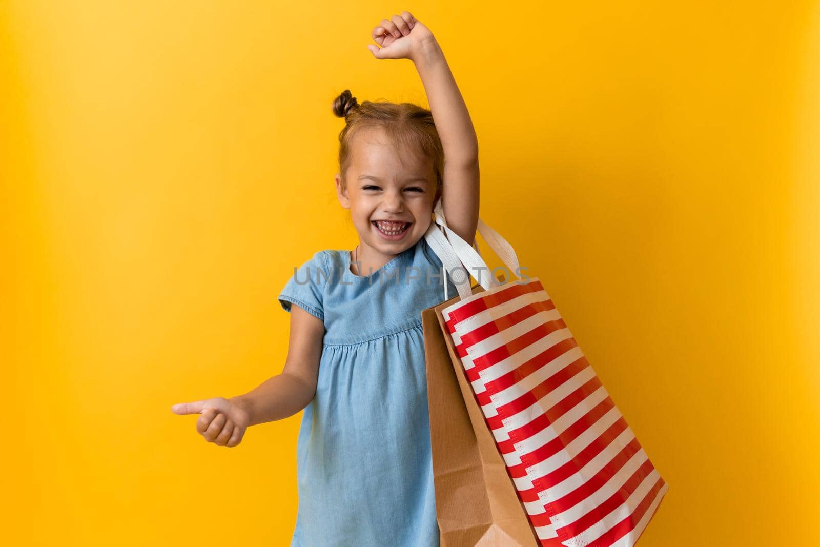 Portrait Caucasian Beautiful Happy Little Preschool Girl Smiling Cheerful And Holding Cardboard Bags Thumb Up Isolated On Orange Yellow Background. Happiness, Consumerism, Sale People shopping Concept by mytrykau