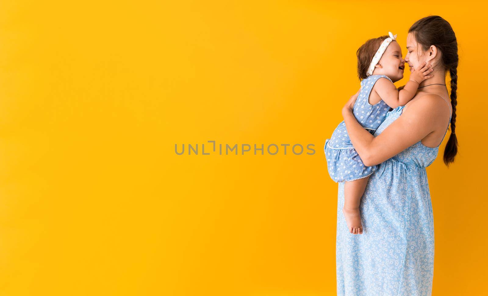 Motherhood, hot summer - Banner young pregnant happy smiling mother woman in blue dress holding little baby daughter toddler sibling in arms looking into eyes kisses on yellow background coppy space.