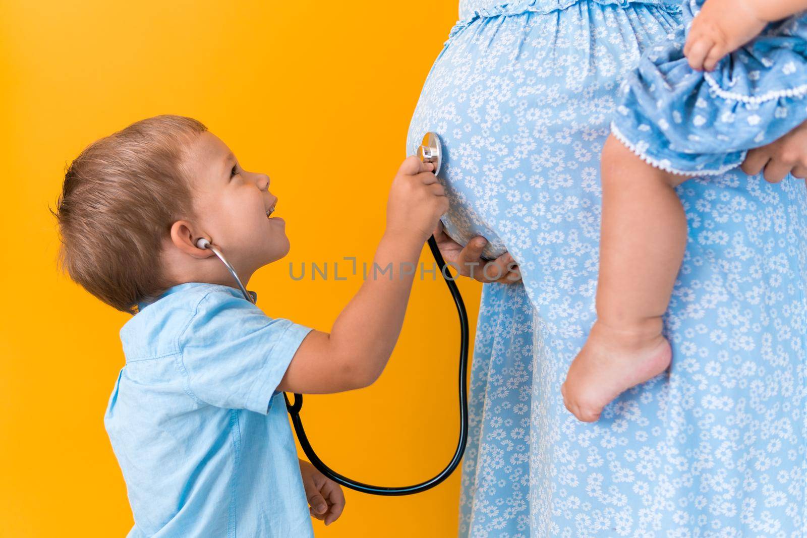 Motherhood, hot summer - croped portrait pregnant mother unrecognizable woman blue dress little boy son sibling treat pregnant mom role play stethoscope unborn brother mom tummy on yellow background.