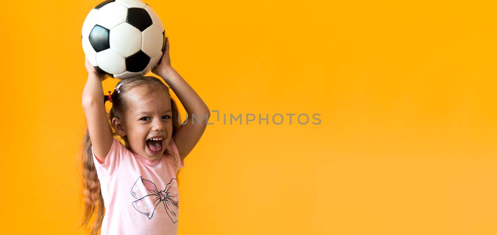 Authentic cute smiling preschool little girl with classic black and white soccer ball look at camera on yellow background. child play football in t-shirt and shorts. Sport, championship, team concept.