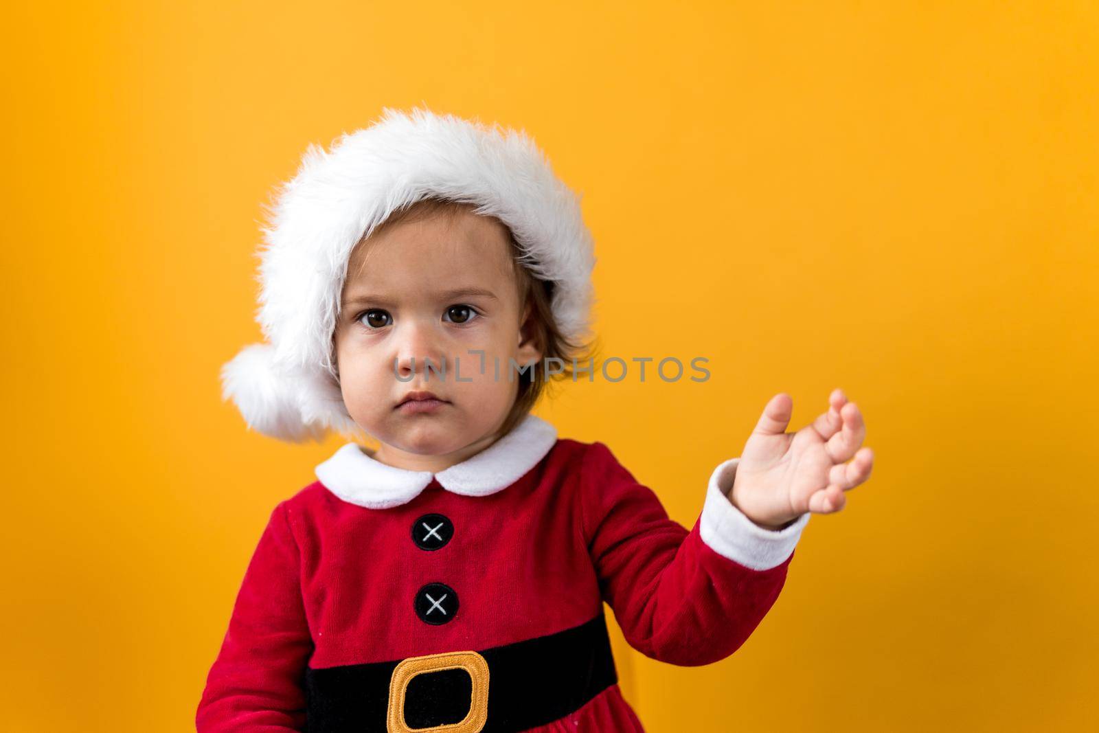 Serious Sad Portraite Cute Happy Cheerful Chubby Baby Girl in Santa Hat Waving Hand At Yellow Background. Child Play Christmas Scene Celebrating Birthday. Kid Have Fun Spend New Year Time Copy Space by mytrykau