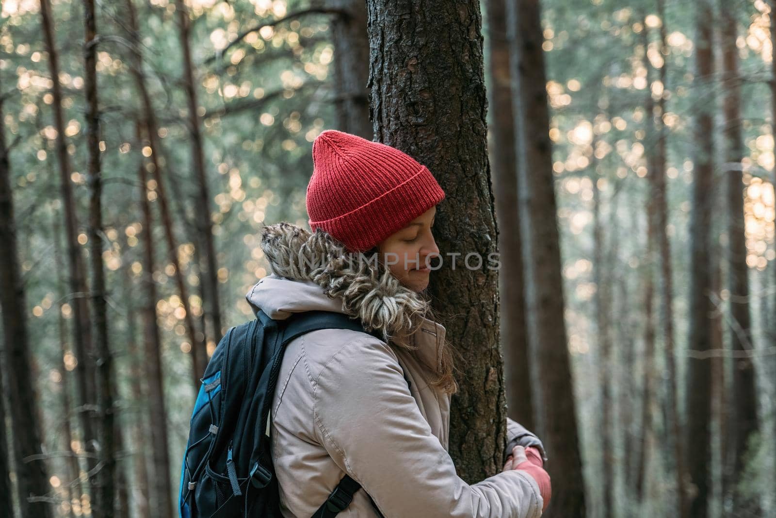Tourist young woman with closed eyes in red cap hugging a tree in the cold forest during the mountain hike. Connecting to nature, protect the environment concept.