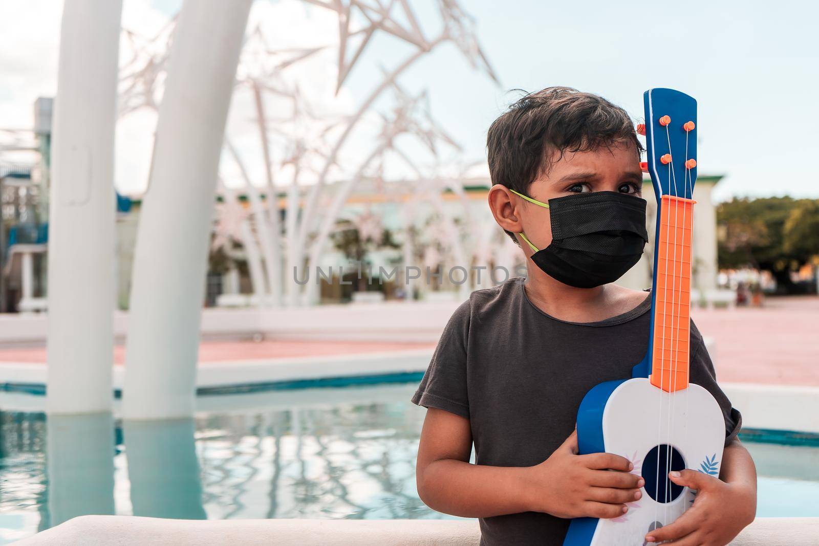 Latin boy outdoors with a medical mask and holding a ukulele in his hands. Concepts of musical training in childhood.