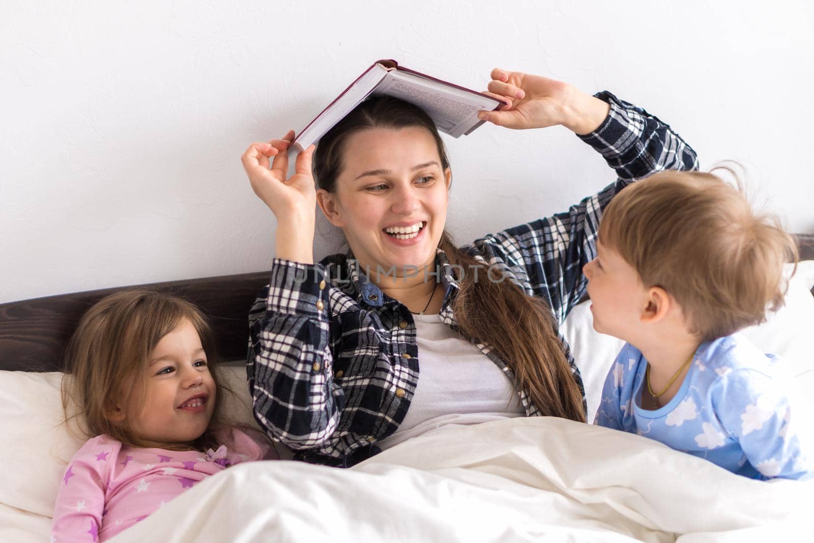 happy family. Close up loving mother lying with daughter son two kids pajamas in bed children reading interesting storybook, preschool smiling kid girl enjoy fairytale before go to sleep Book on head.