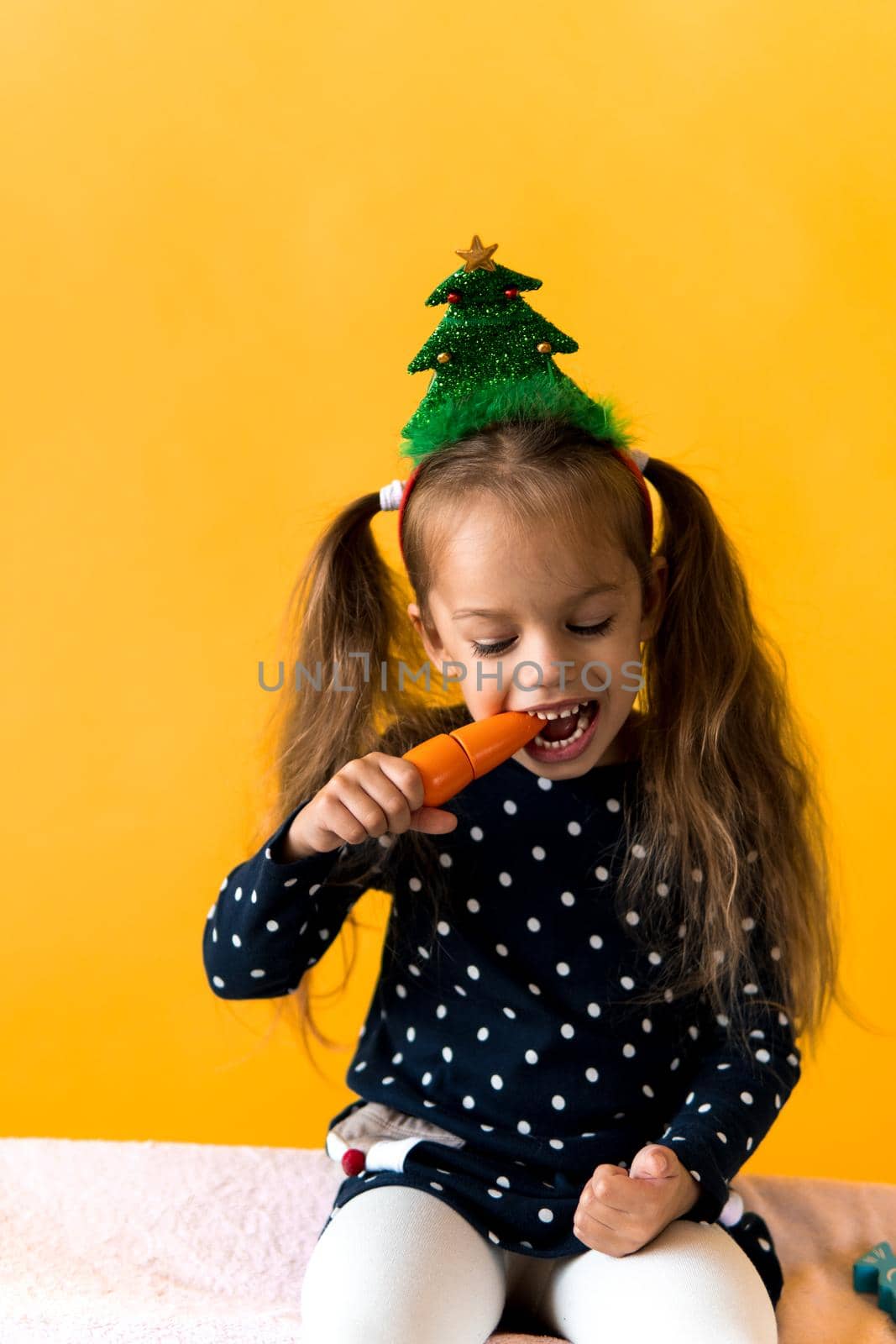 Portrait positive cheerful smiling happy little schoolgirl girl Christmas tree decoration polka dot dress biting eat orange carrots on orange background. New year, holiday, celebration, winter concept.