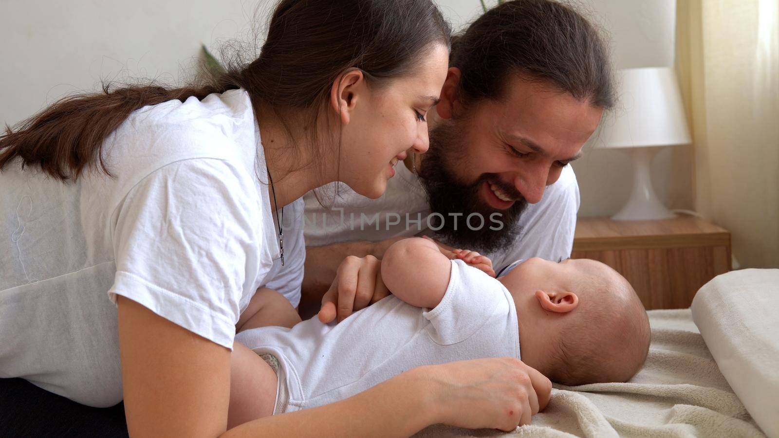 Woman And Man Holding Newborn. Mom, Dad And Baby On Bed. Close-up. Portrait of Young Smiling Family With Newborn On Hands. Happy Marriage Couple On Background. Childhood, Parenthood, Infants Concept.