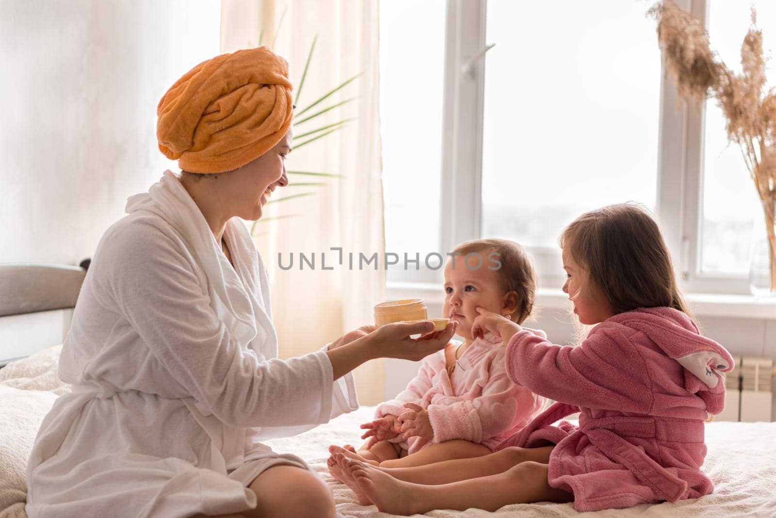 Young mother two daughters in curlers bathrobes. happy smiling family skin care concept. Mom teaches little kids child to do makeup smear cream on face sittting in orange bathrobes on bed in bedroom by mytrykau