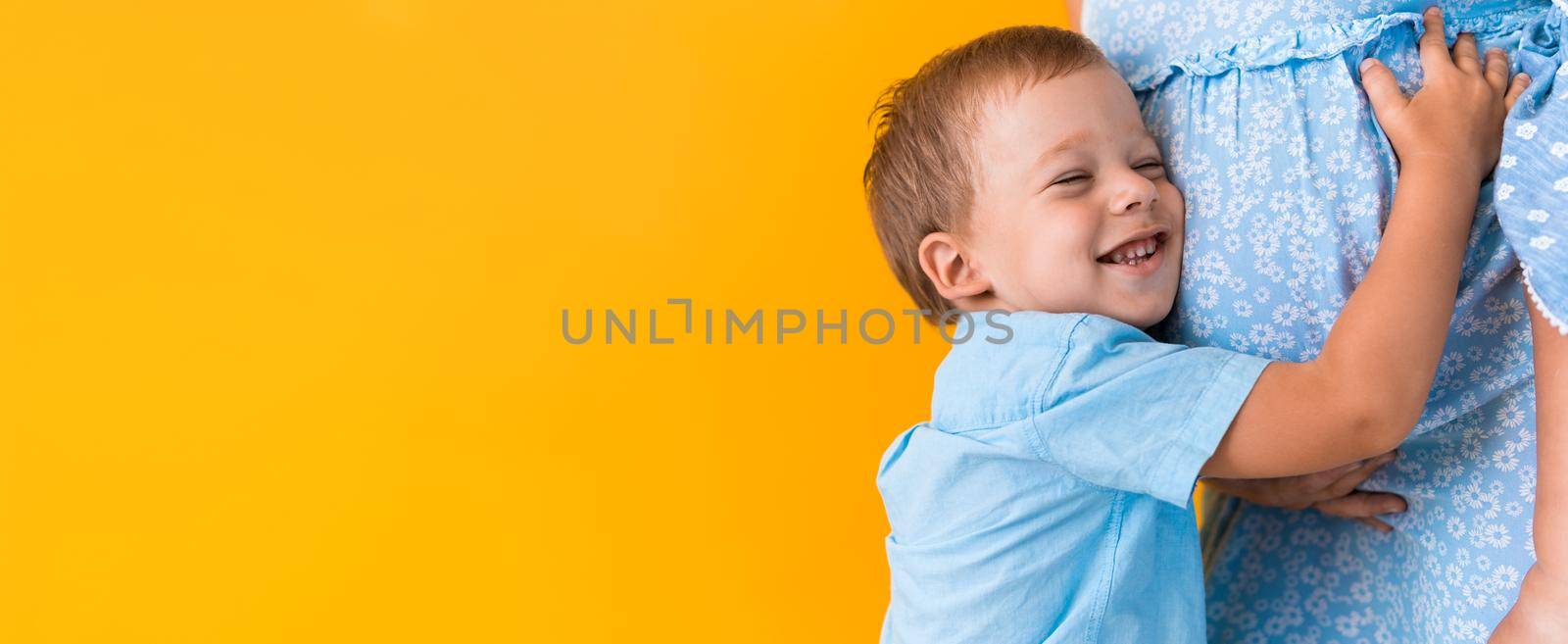 Motherhood, femininity, love, Childhood, hot summer - croped banner pregnant unrecognizable woman blue dress little preschool boy son sibling hug mum big belly brother on yellow background copy space.