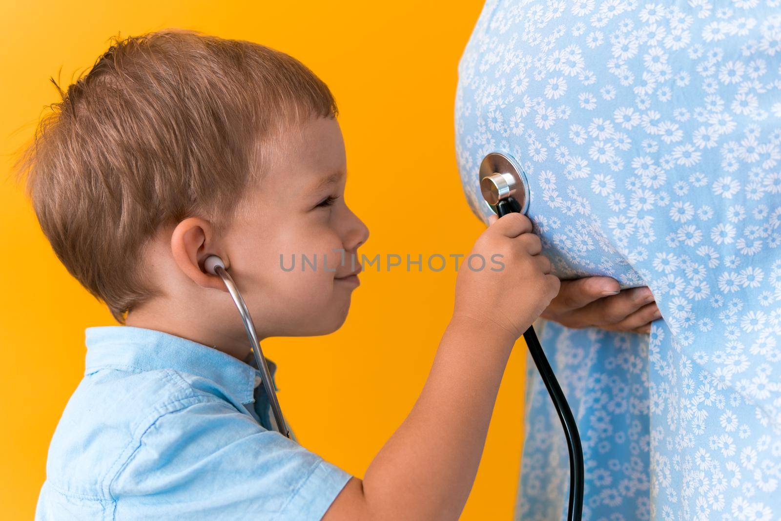 Motherhood, hot summer - croped portrait pregnant mother unrecognizable woman blue dress little boy son sibling treat pregnant mom role play stethoscope unborn brother mom tummy on yellow background.