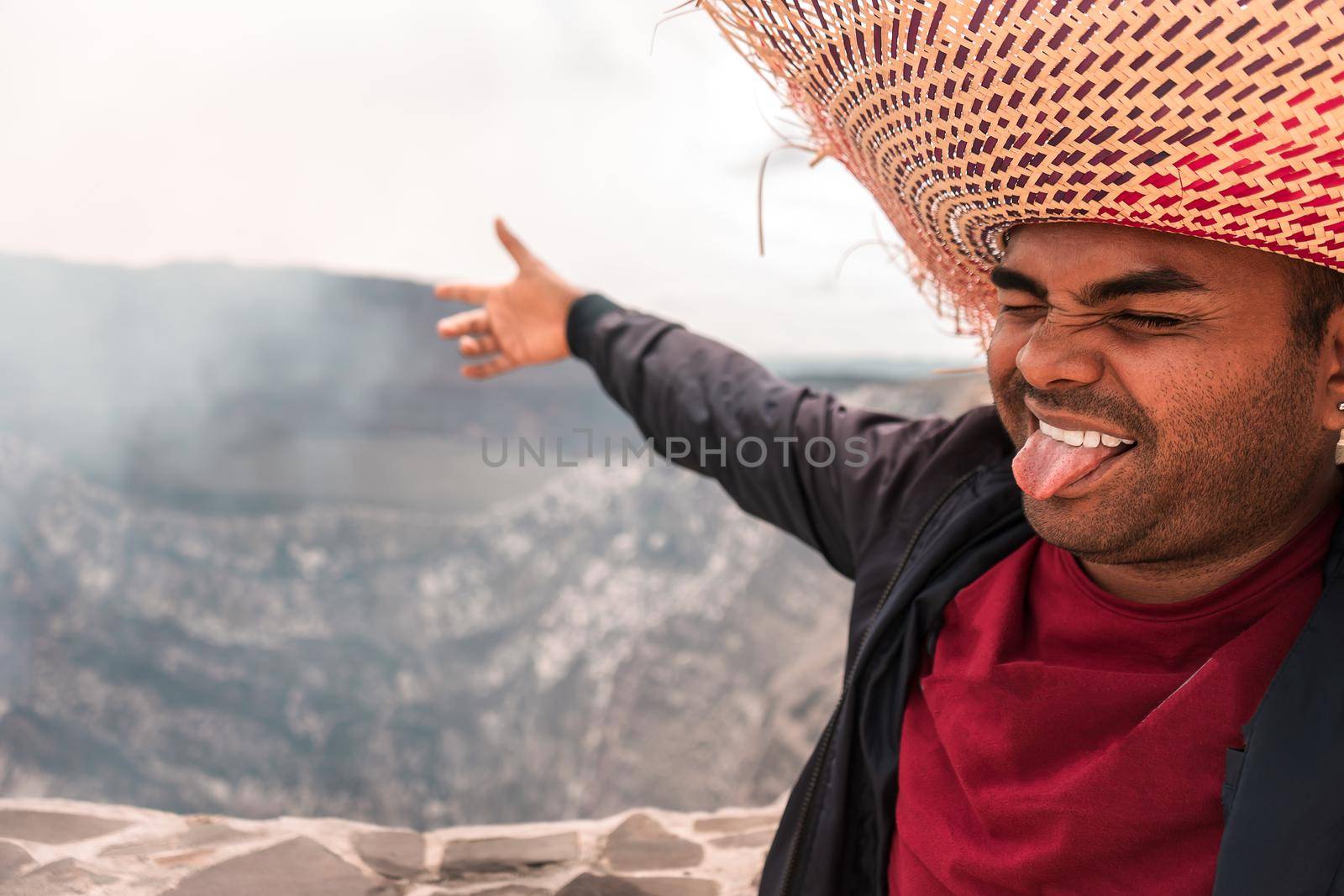 Latin man with a small three-day beard wearing a striking hat and sticking out his tongue pointing towards the crater of a volcano during an adventure trip in Masaya, Nicaragua.