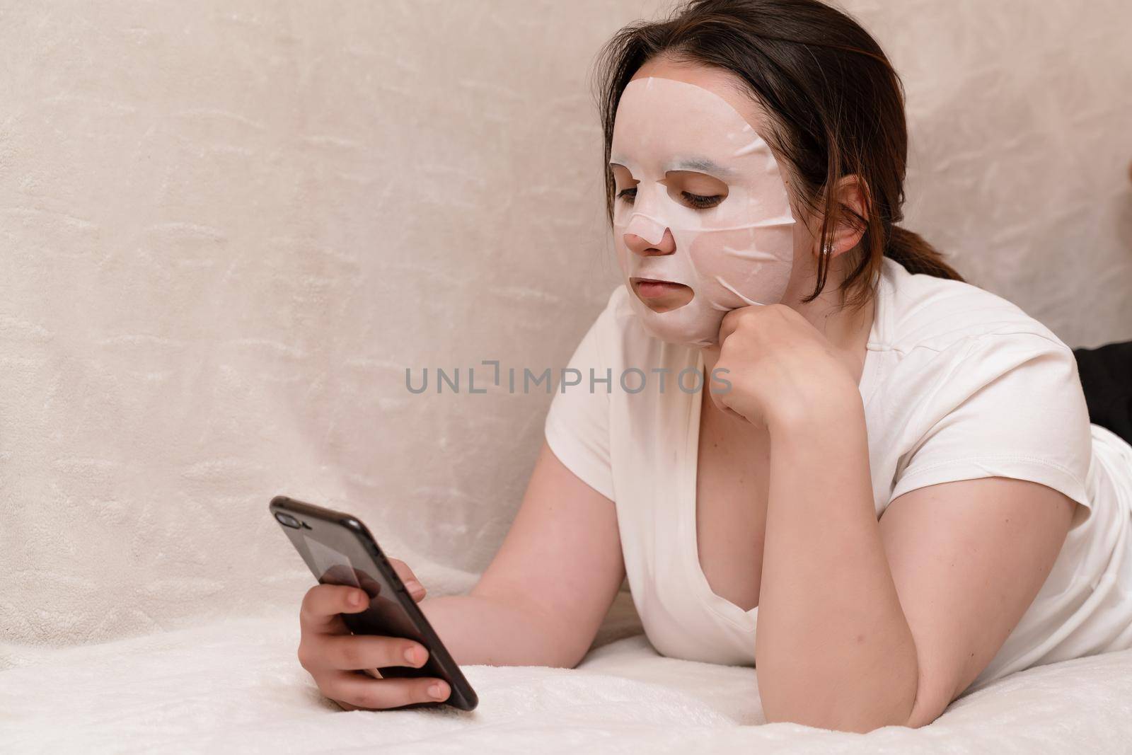 A young woman in a mask of cosmetic fabric lies on a sofa in a white T-shirt and surfs in a smartphone. The concept of quarantine, home care yourself