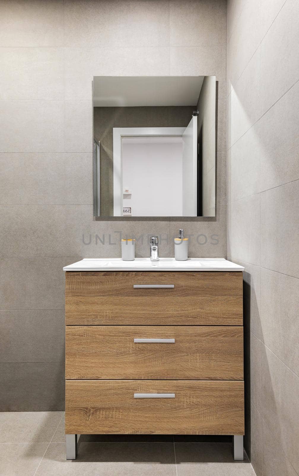 Modern refurbished bathroom in minimalist style with gray tiled walls, mirror and wooden drawers under sink