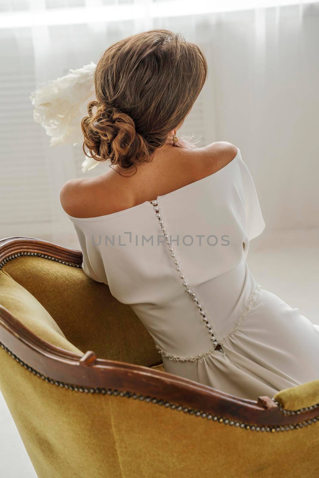 A beautiful girl in a white dress is sitting back in a chair. The back of the dress is decorated with buttons, a beautiful neckline, hair is gathered in a beautiful bun. An antique chair brown tones