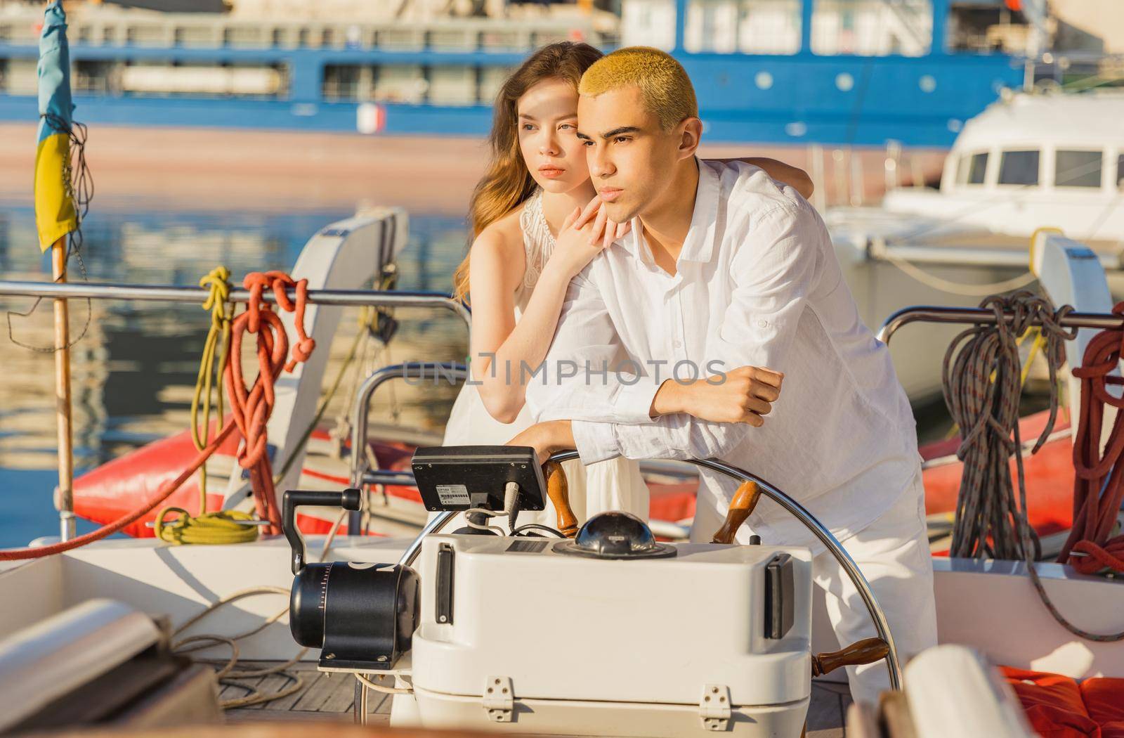 a couple in white clothes stands near the steering wheel on a yacht