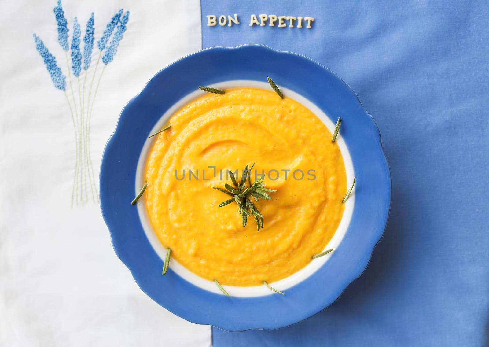 pumpkin soup in a blue plate with rosemary on a napkin with flowers, lettering bon appetit