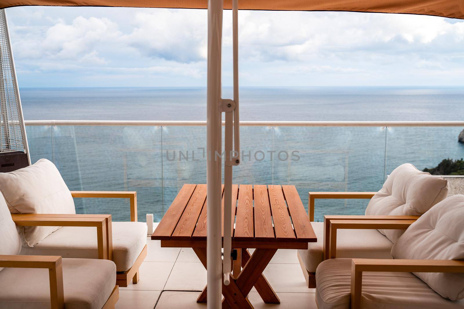 Restaurant with sea view. You can see rocks in the sea. Wooden tables and chairs with white cushions. Overcast, cloudy weather. by Matiunina