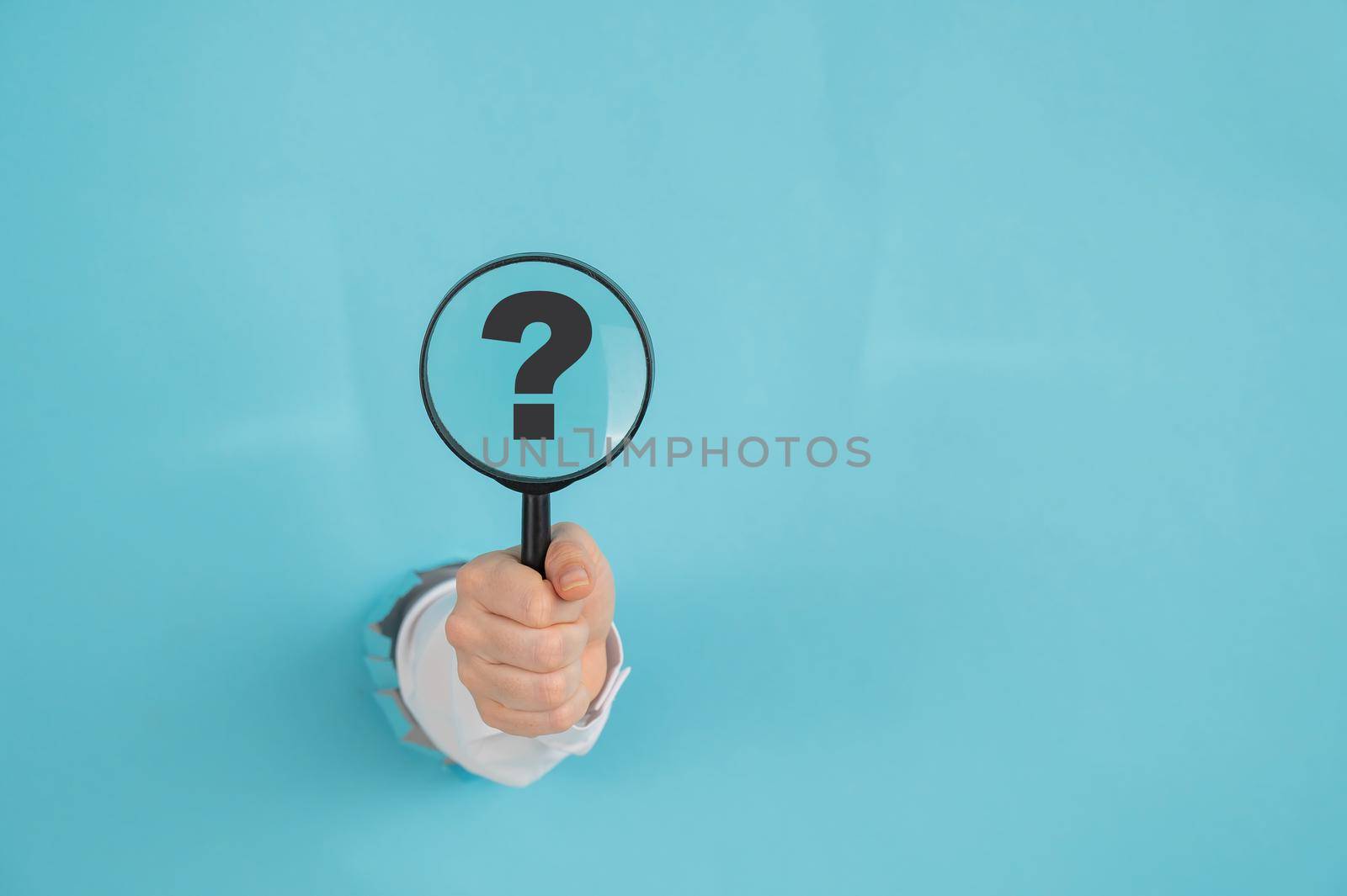 A woman's hand sticking out of a hole on a blue paper background holds a magnifying loupe with a question mark