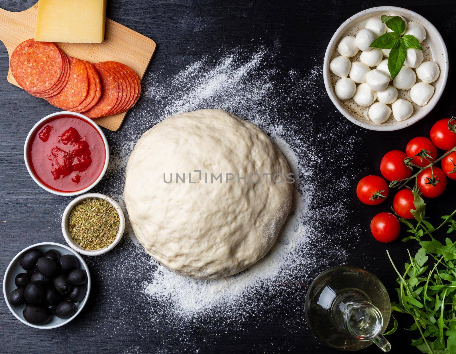 Raw dough for pizza with ingredients and spices on dark black background, top view, flat lay.