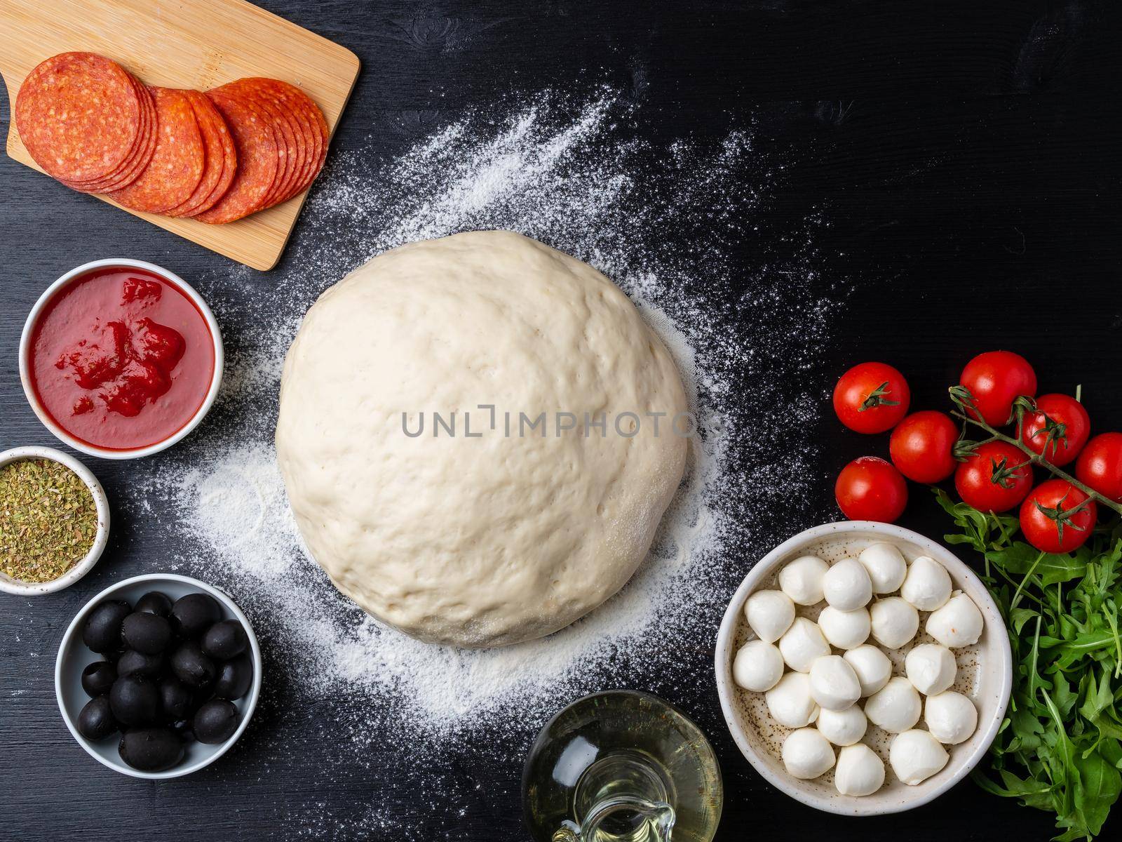 Raw dough for pizza with ingredients and spices on dark black background, top view, flat lay, copy space.