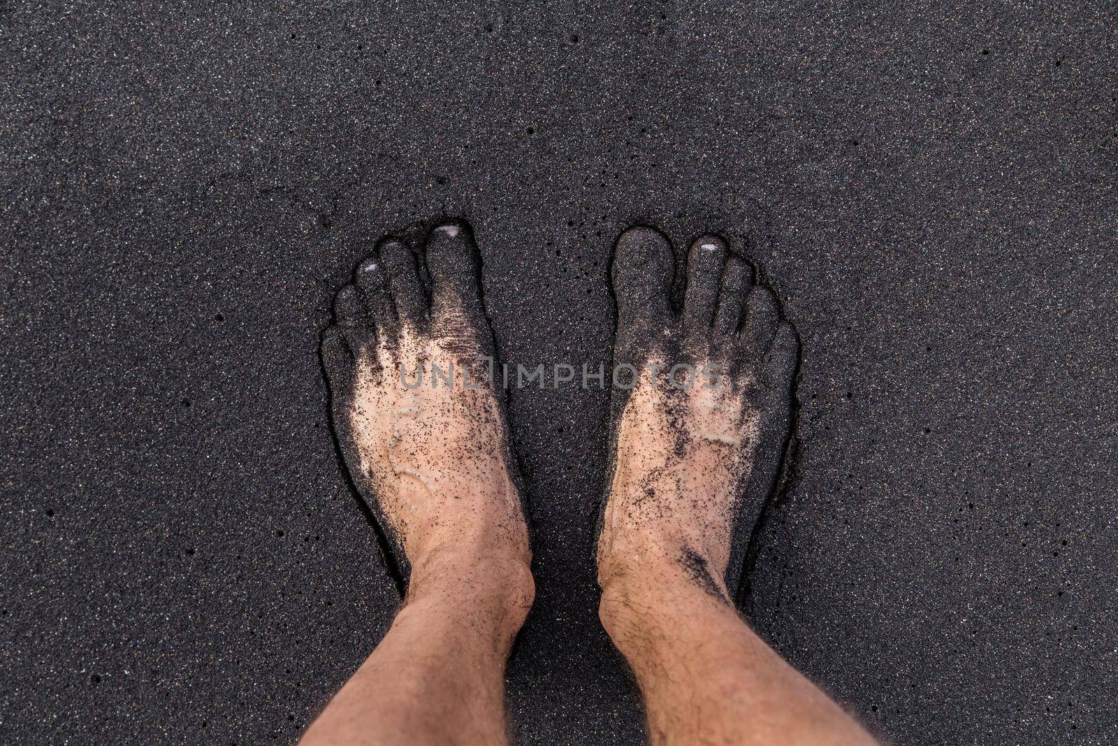 A man standing barefoot on volcanic black sand on the Tenerife beach, Canary Islands. Two feet covered by the sand.