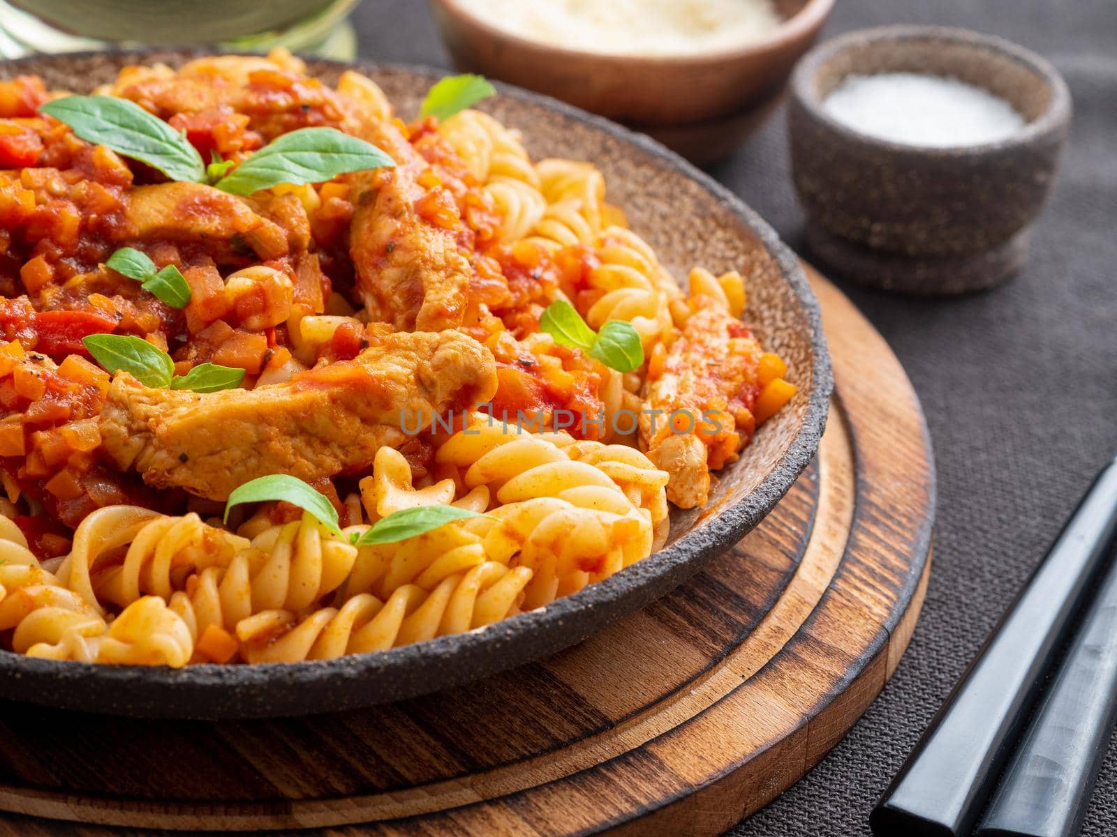 fusilli pasta with tomato sauce, chicken fillet with basil leaves on dark brown background, side view.