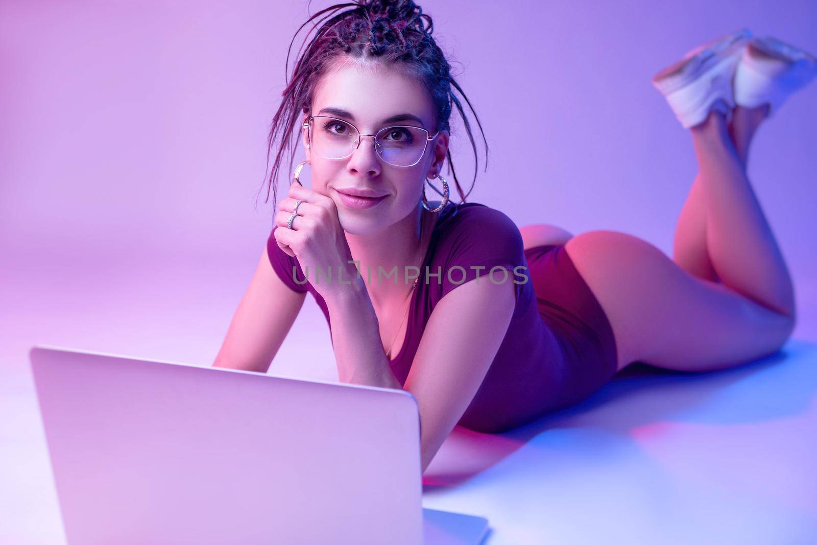 the a slender girl on a white background in neon color is lying on the floor looking at a laptop. learn exercises and training online