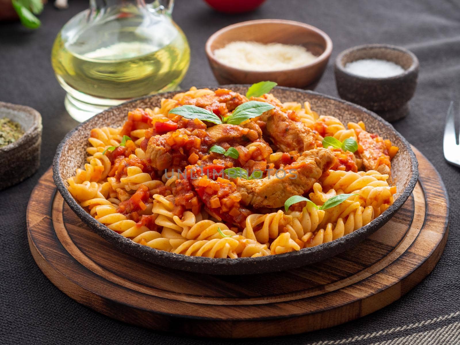 fusilli pasta with tomato sauce, chicken fillet with basil leaves on dark brown background, side view.