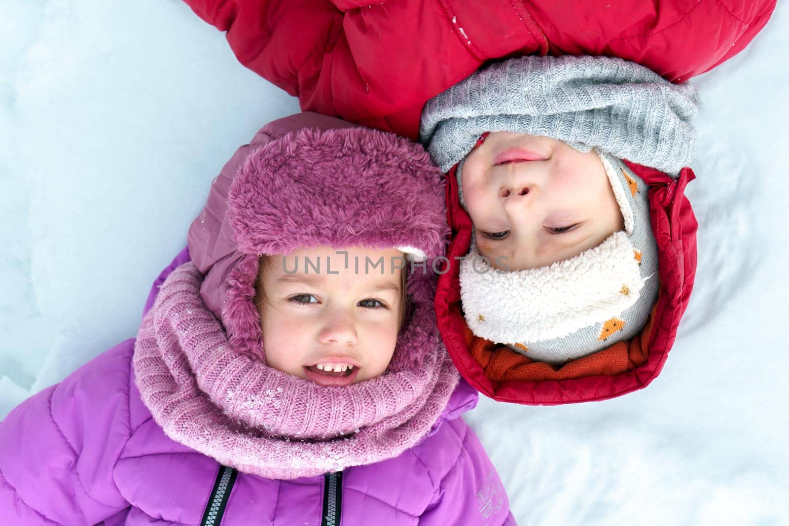 Childhood, snowy time, winter holidays cold weather. Close up portrait little preschool happy Children boy girl face, Smilling kids in warm knitted hat and scarf laying down on white snow background by mytrykau