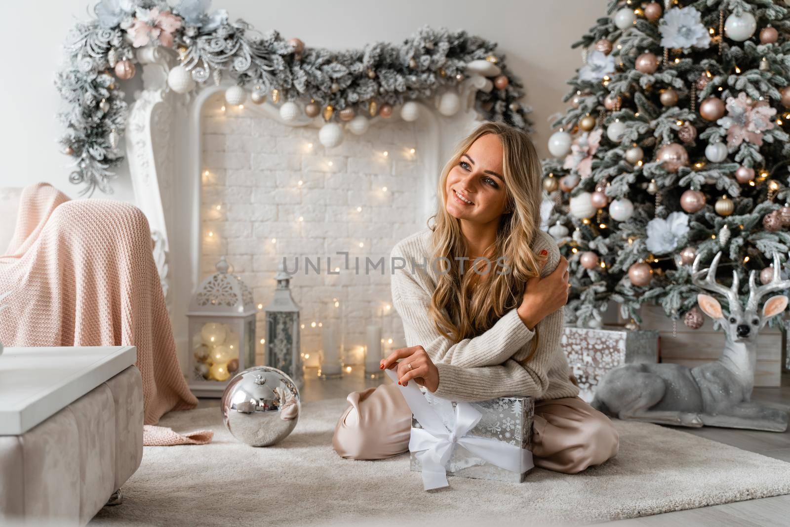 Happy blonde beauty enjoys Christmas Under the Christmas tree in a cozy home, holding a box with a gift