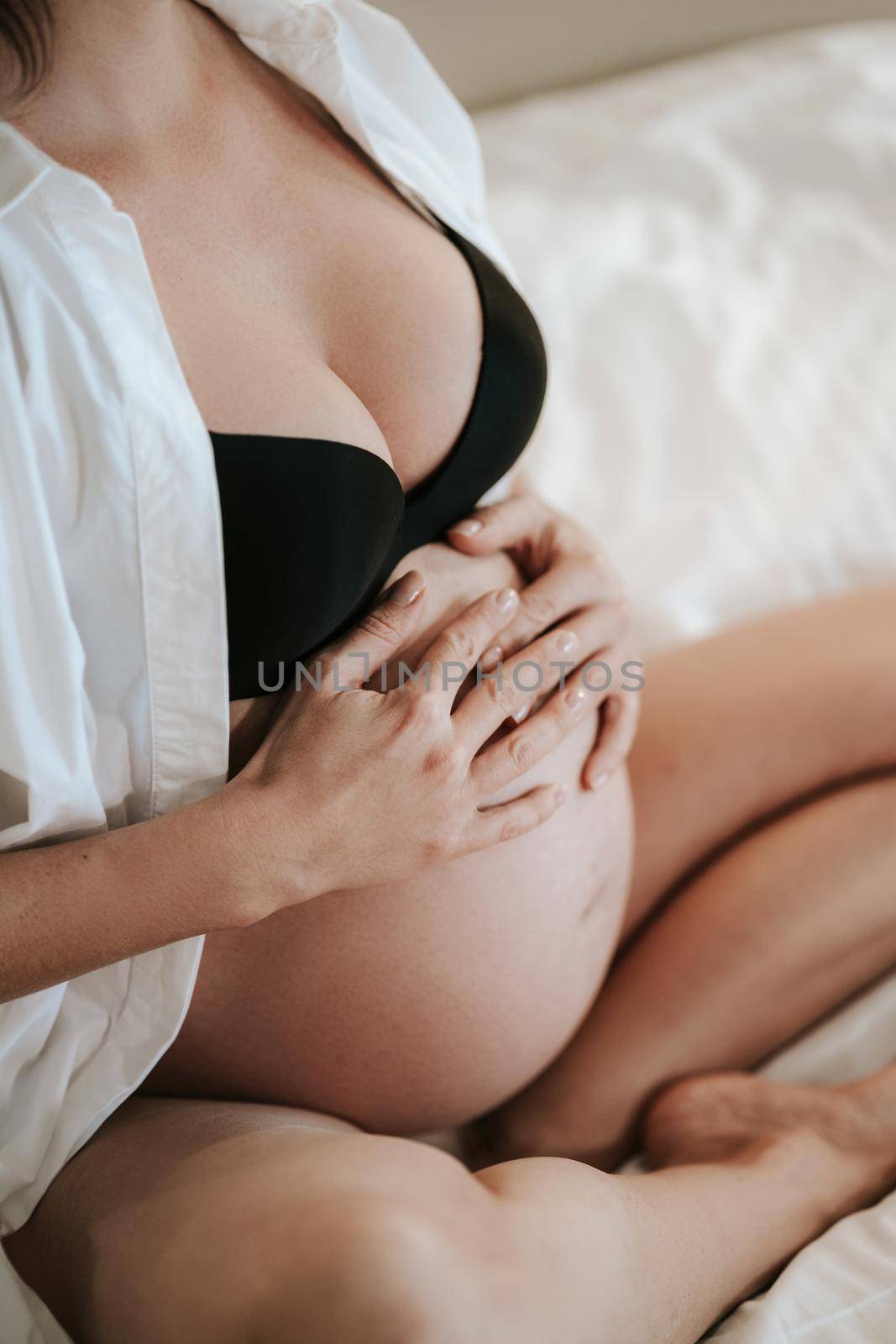 Pregnant woman sits on bed in black bra holding hands on belly. big belly. High quality photo