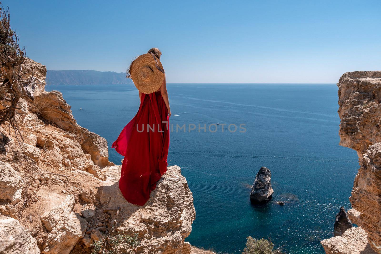 A girl with loose hair in a long red dress descends the stairs between the yellow rocks overlooking the sea. A rock can be seen in the sea. Sunny path on the sea from the rising sun by Matiunina