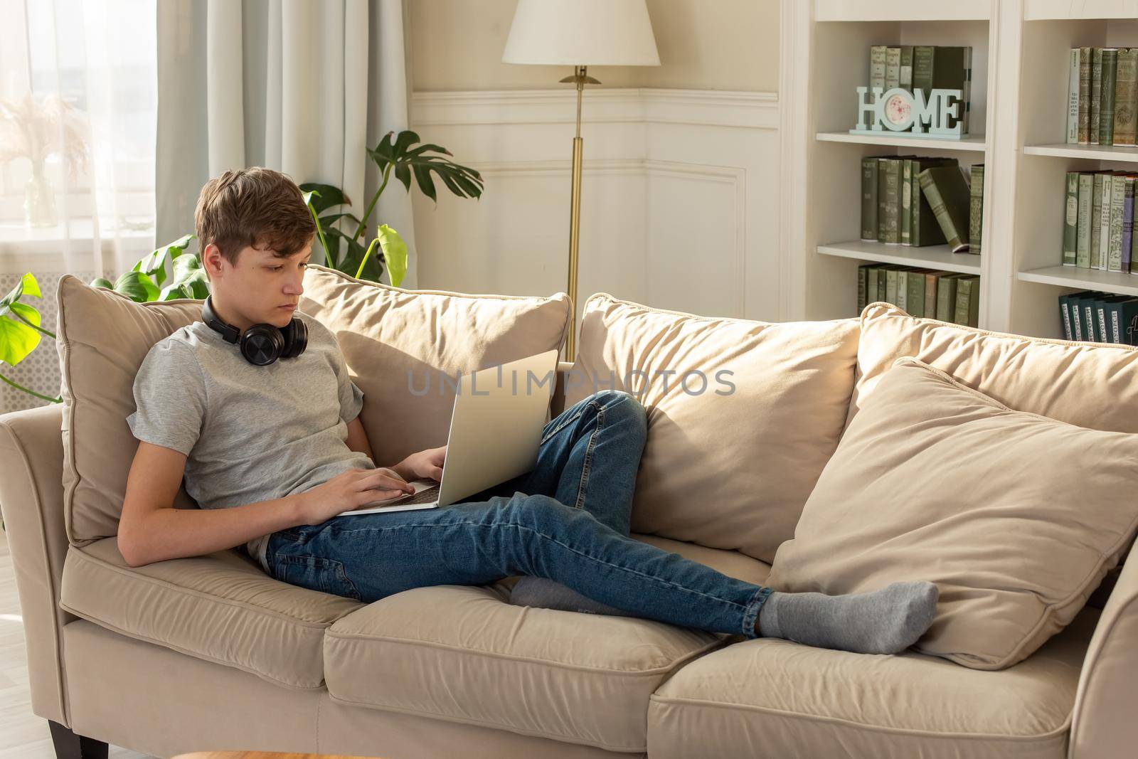 A teenager boy in a gray T-shirt and blue jeans, sits on a beige sofa, in room with plant, wearing black headphones around his neck, looks into a laptop. Copy space