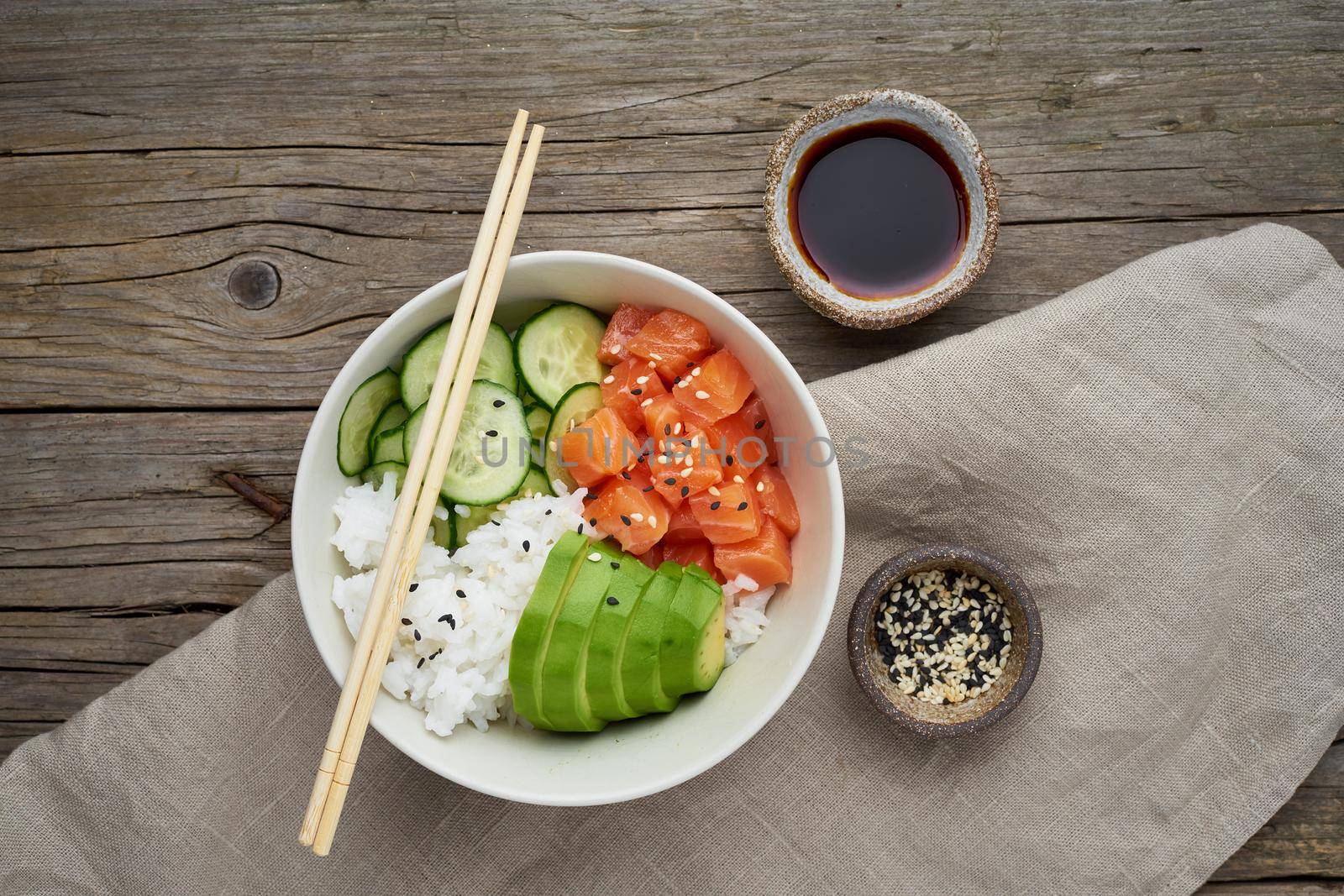 Salmon poke bowl with fresh fish, rice, cucumber, avocado with black and white sesame. Old wooden table. Food concept.